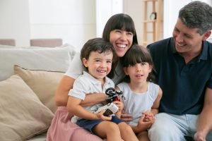 Cheerful funny parents and two kids watching funny movie at home, sitting on couch in living room and looking away and laughing. Medium shot. Leisure time with family or television concept